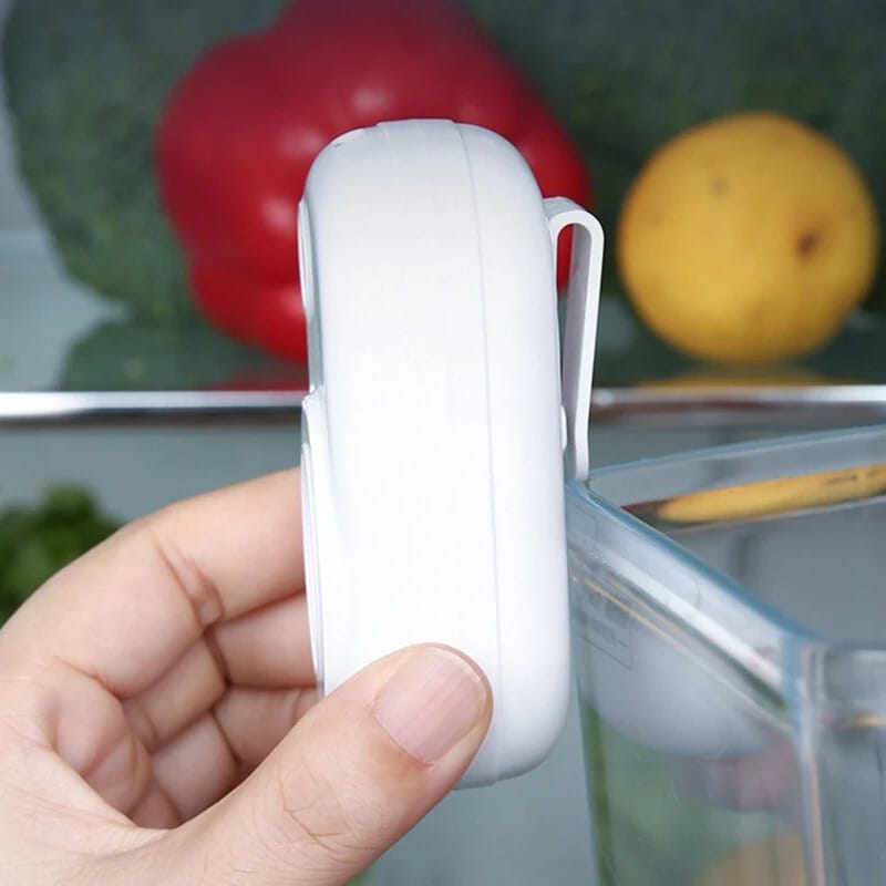 Mano che regge il cubo di lato mostrando la clip di aggancio, davanti a un peperone e a un limone in frigo