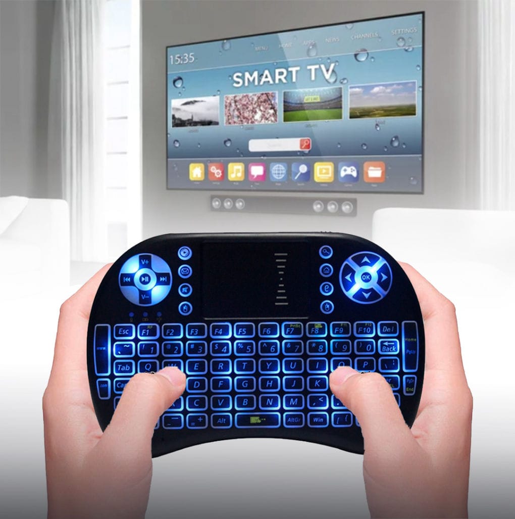 Due mani impugnano la mini tastiera retroilluminata di blu davanti a una Smart TV accesa in salotto
